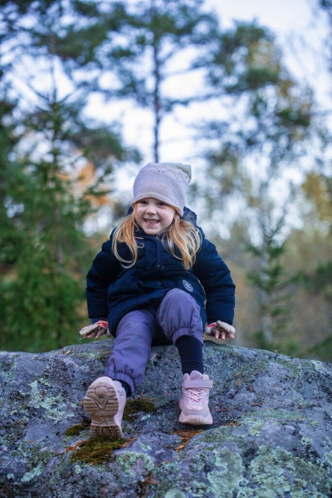 A smiling young girl sits on a rock outdoors in winter clothing, enjoying a day in nature.